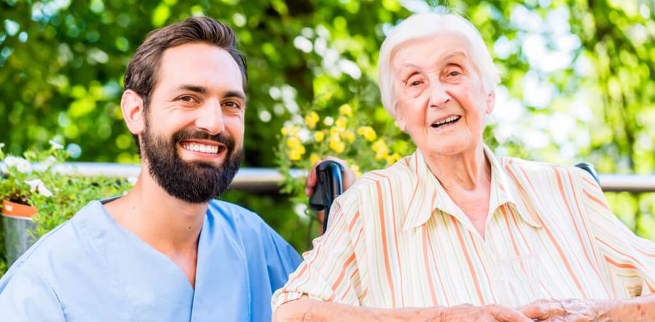 Männliche Pflegekraft und Seniorin im Rollstuhl lächeln in die Kamera