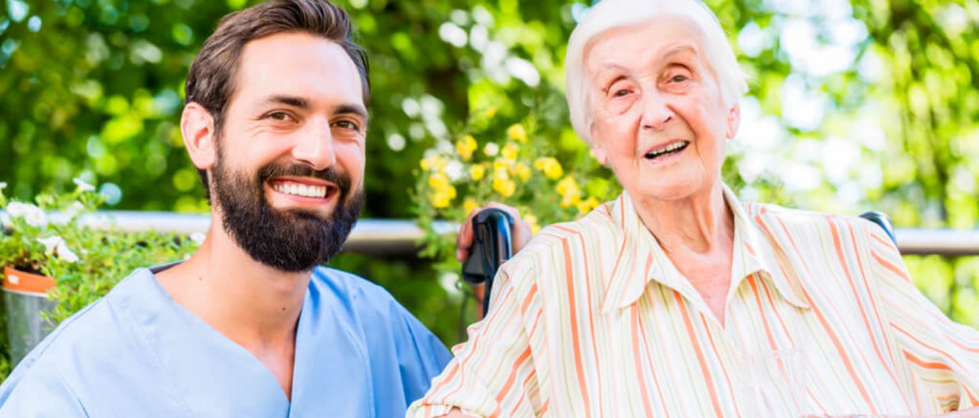 Männliche Pflegekraft und Seniorin im Rollstuhl lächeln in die Kamera