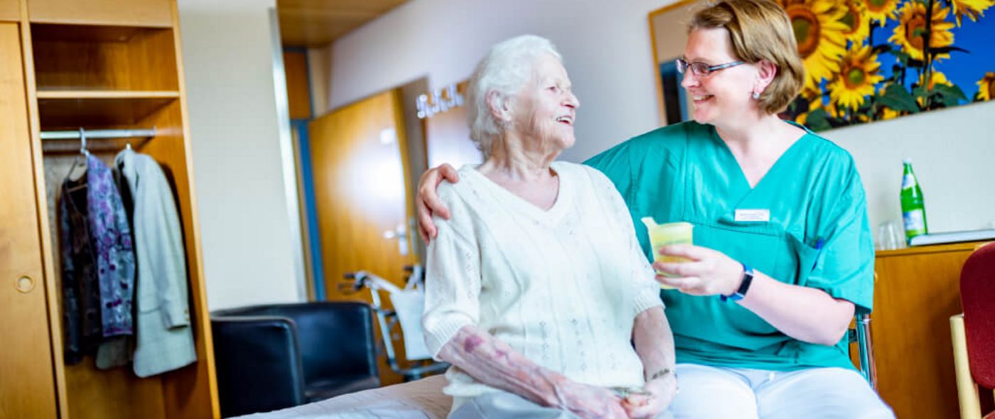 Pflegekraft reicht Seniorin einen Becher mit Saft, während Sie auf dem Bett sitzen