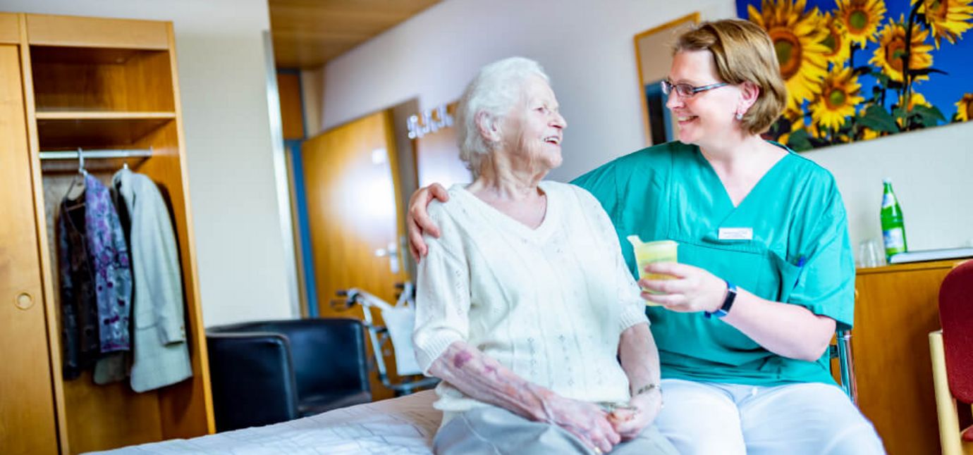 Pflegekraft reicht Seniorin einen Becher mit Saft, während Sie auf dem Bett sitzen